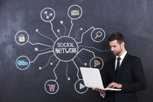 Social networking. Handsome young man working on laptop while standing against social network chalk drawing on blackboard